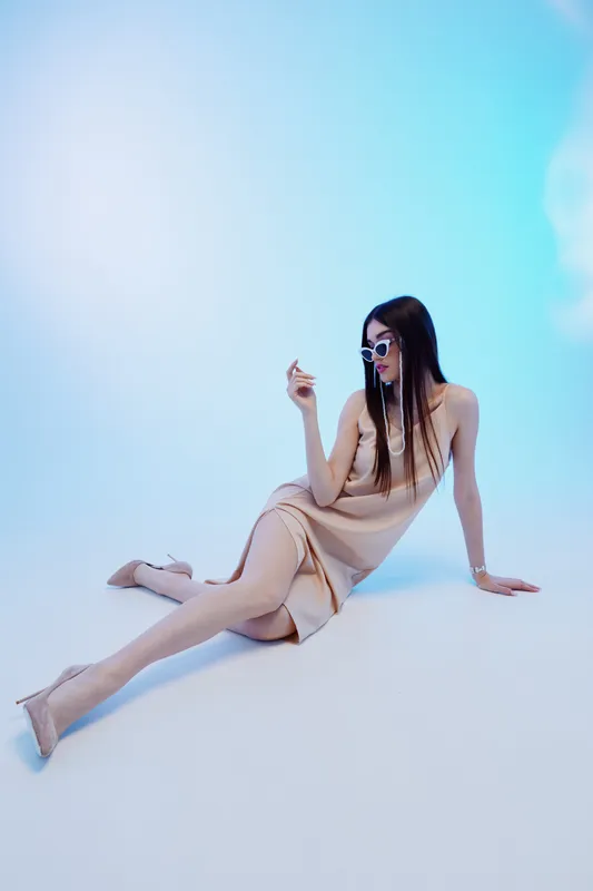 Elegant woman in a beige dress poses sitting on the floor wearing sunglasses. Studio shot on a light blue background. Concept of beauty, fashion, and style.
