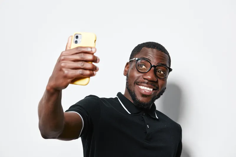 African American man with dark skin and glasses taking selfie with smartphone smiling in casual black polo shirt on white background indoors.