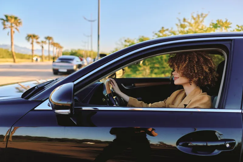 A woman drives a sleek car on a sunlit road, captured with retro film color tones. A candid lifestyle moment highlighting independence, travel, and everyday journeys.