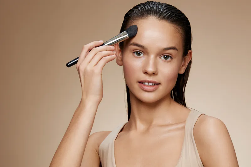 Young woman applying makeup with a brush, showcasing natural beauty and confidence against a warm beige background for beauty and skincare concepts