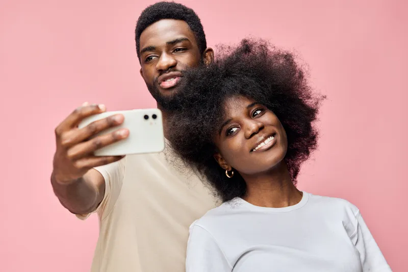 A joyful young African couple taking a selfie together on a pink background, showcasing natural beauty and happiness in casual clothing