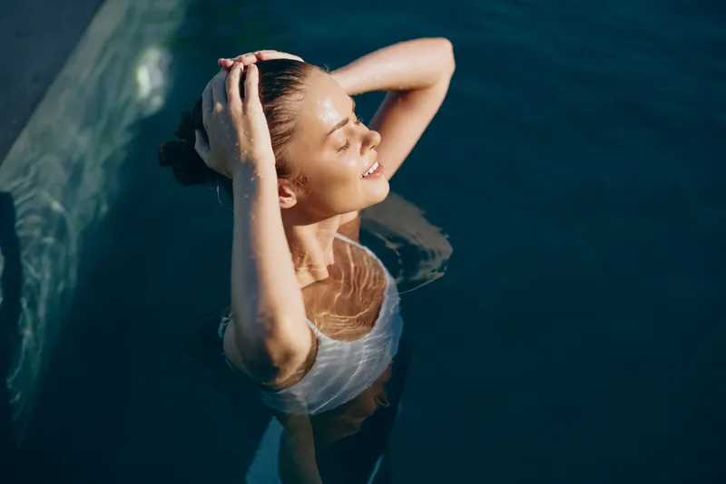 Relaxed woman in a sunlit pool, head back, hands in her hair, wearing a white swimsuit, enjoying warm weather, calm water, serene leisure portrait, outdoor wellness moment
