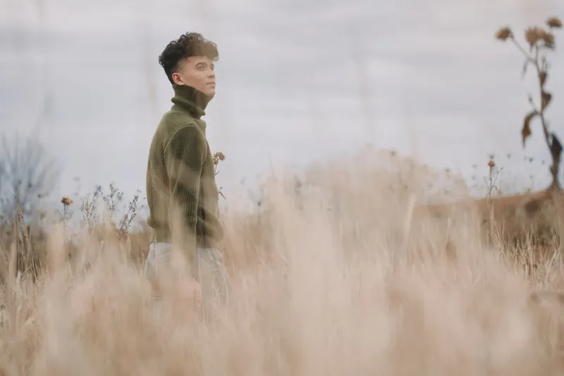 Candid portrait of a young man standing in a windswept field wearing a green sweater, captured in natural light to convey a genuine, unposed moment with tactile grasses and quiet presence.