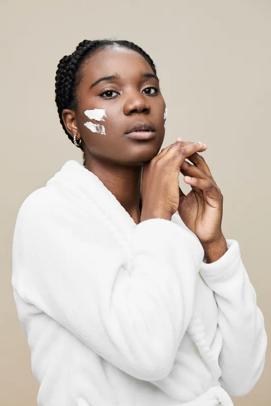 Young woman with natural hair applying skincare in a bathrobe, showcasing a calm expression against a neutral background, reflecting self care and beauty