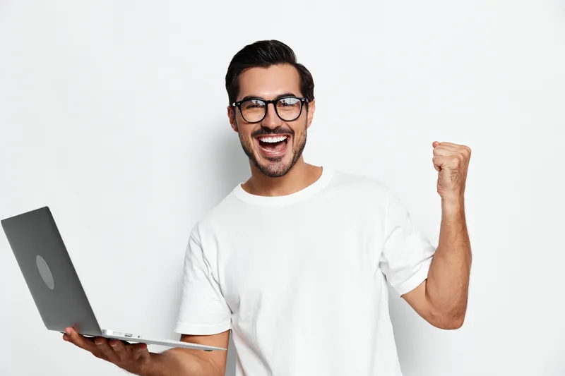 Excited young caucasian man wearing glasses holding laptop celebrating success with raised fist, looking at camera, isolated on plain white background. Work achievement concept