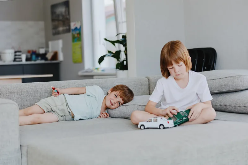 Two boys playing on a cozy sofa, one is lying and smiling while the other is focused on his toy car. A bright and inviting indoor setting enhances the playful atmosphere.