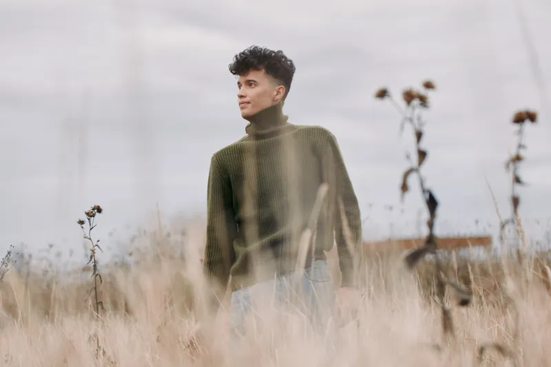 Young man stands in a windswept meadow wearing a green turtleneck, shot in natural soft light to convey authenticity and credibility through honest, candid portraiture.