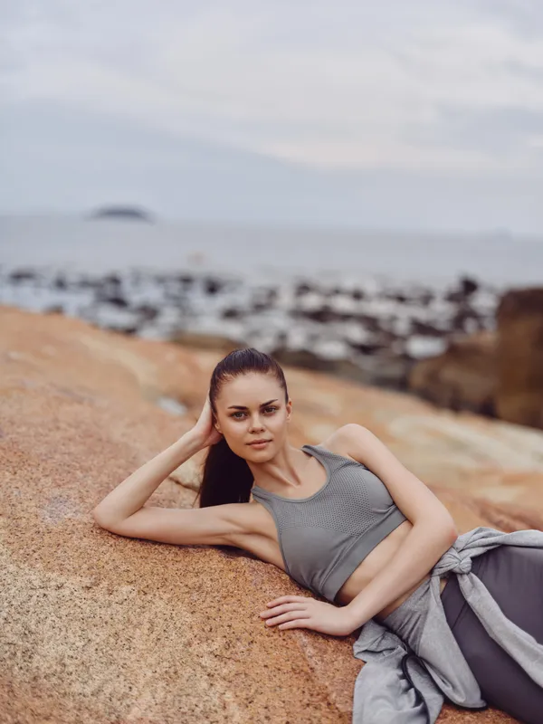 Young woman in sporty attire lounging on rocky beach, showcasing a healthy lifestyle and serene emotions with a coastal background and muted color palette