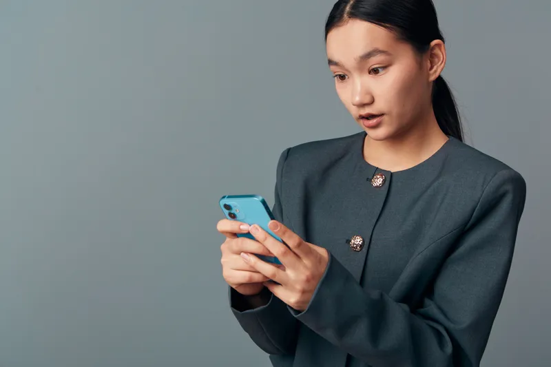 Woman holding smartphone with both hands looking surprised or shocked with mouth slightly open against neutral gray background in casual formal clothing for technology or reaction