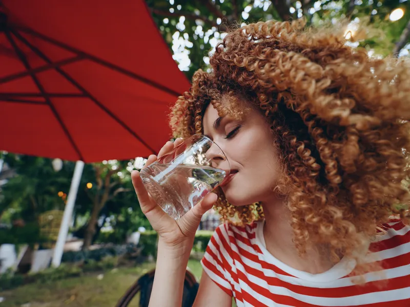 A woman with curly hair drinks water from a glass outdoors under a bold red parasol. She wears a striped shirt, capturing a fresh, casual moment of hydration in sunlight.