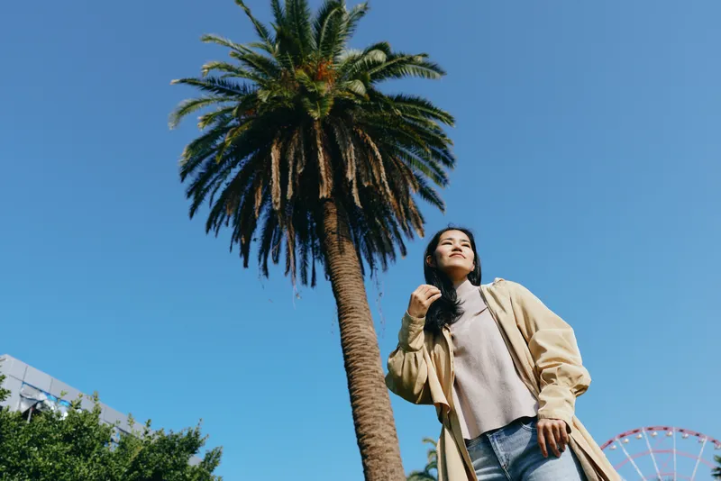 Low angle view of a casually dressed figure beside a tall palm tree under a clear blue sky, capturing urban greenery and sunny street style with a relaxed jacket.