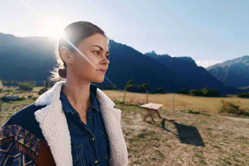 Woman portrait mountains sunlight nature meadow jacket travel. Young woman in denim shirt and shearling jacket stands in an alpine meadow with warm backlight, looking thoughtfully toward distant