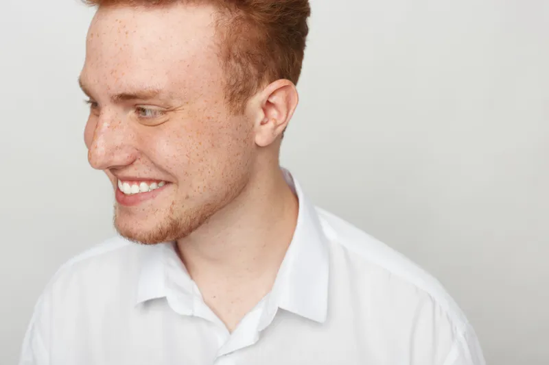 Smiling young man with red hair and freckles looks to the side wearing a white shirt. Studio portrait on a light background with natural expression and casual style.