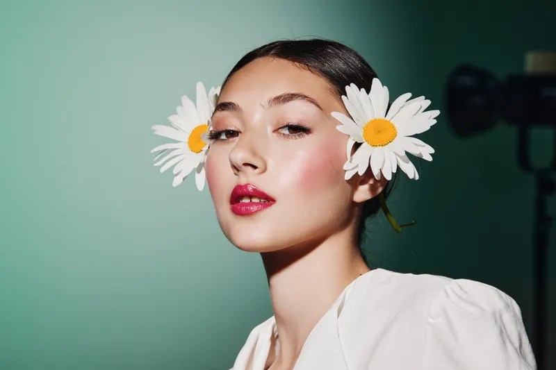Close-up portrait of young woman with glossy makeup wearing large white daisy flowers near her ears. Soft studio lighting highlights fashion beauty concept on green background.