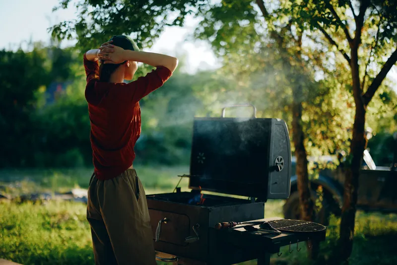 A young woman cooking on a barbecue grill outdoors in a green park, enjoying a sunny day with a relaxed expression, perfect for summer gatherings