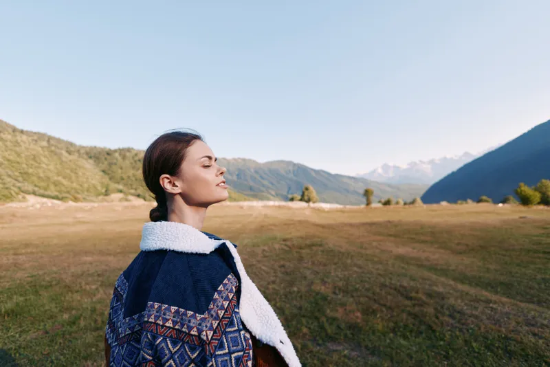 Woman portrait in mountains meadow outdoors, peaceful moment with closed eyes and cozy patterned jacket. Travel lifestyle, nature scenery, fresh air and mindful relaxation.