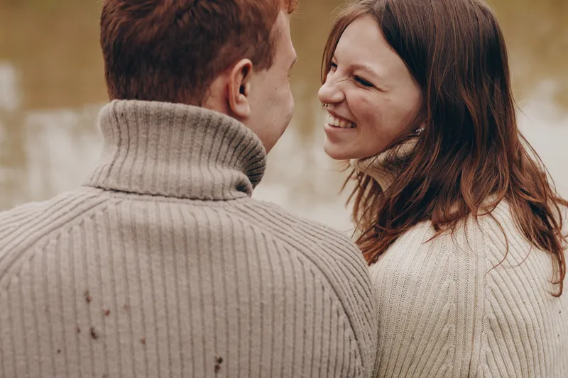 Joyful couple outdoors, smiling at each other, warm sweaters, sunlit moment, relaxed posture, soft breeze, natural colors, autumn backdrop, candid affection, close connection.