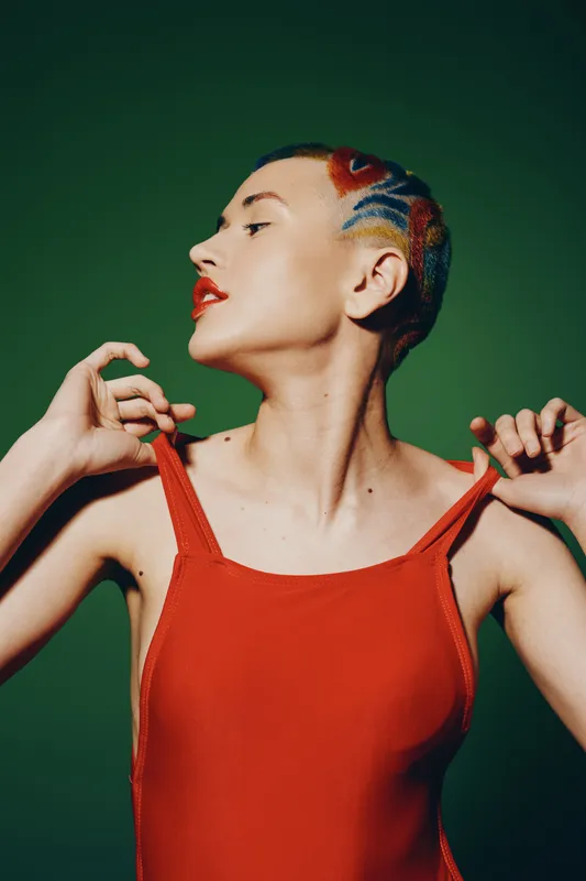 Artistic woman with short colorful hair wearing a red sleeveless top poses confidently against a solid green background with bold, modern style.