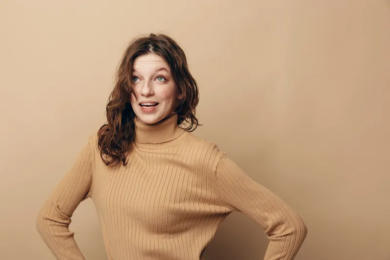 Young woman with curly hair wearing beige sweater looks surprised and happy with hands on hips. Studio portrait on neutral background. Concept of emotion, expression, and casual style.