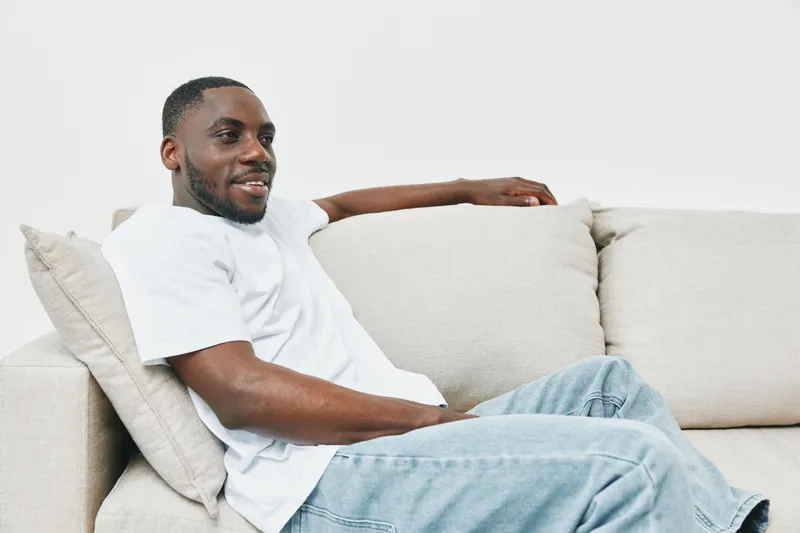 Smiling African American man relaxing on a beige couch in casual white apparel, showcasing comfort and style, ideal for lifestyle and happiness themes