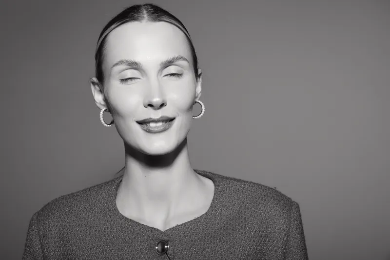 Closeup of a young white woman with fair skin and slicked back hair smiling with closed eyes wearing hoop earrings and a textured sweater against a plain background.