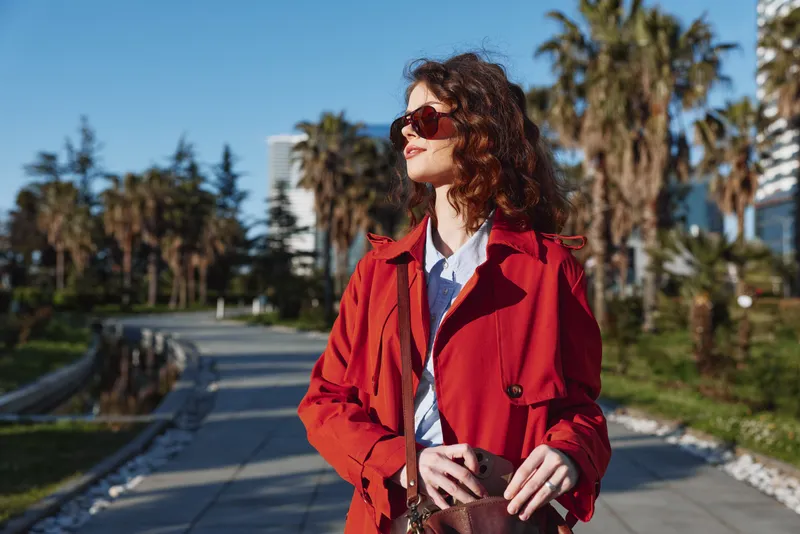 Stylish woman in a red coat walks along a sunny promenade, wearing sunglasses and a crossbody bag, expressing confidence and casual chic