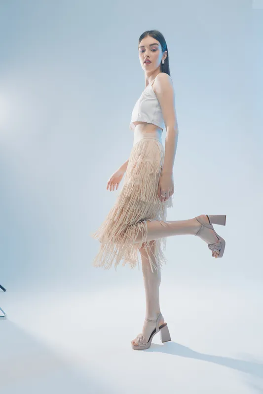 Elegant woman in a white crop top and beige fringe skirt poses lifting one leg in a bright studio. Concept for beauty, fashion, and modern style.
