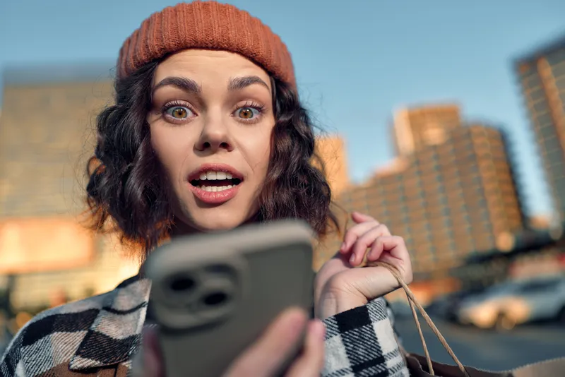 Woman smartphone surprise shopping city portrait expression beanie candid portrait at golden hour glow, capturing authenticity, emotional storytelling and mindful living in urban moment.