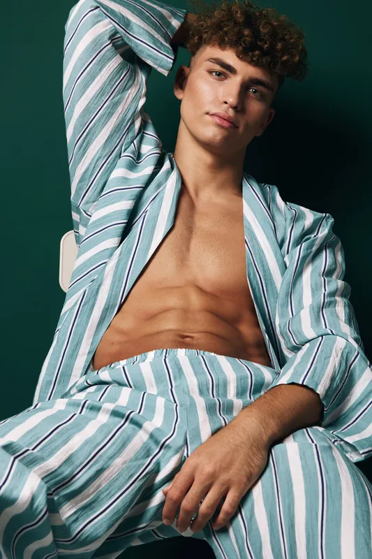 Young man with curly hair wearing striped open shirt and pants sitting relaxed on chair with hand behind head against dark background in casual fashion style portrait.