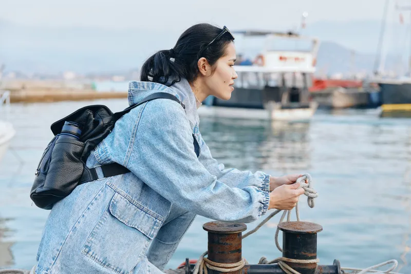 Harbor scene with a woman in a denim jacket and backpack tying a rope to a bollard, boats and calm water in the background, outdoor maritime activity and travel preparation.