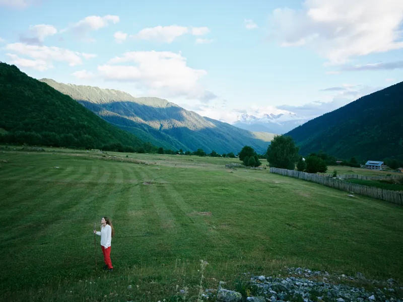 Woman in white top and red pants walks across a green field, distant mountains rise under a blue sky, capturing peaceful rural scenery and wide open space.