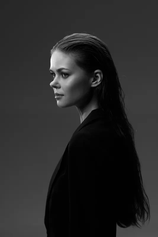 Black and white portrait of a serious woman with long hair in profile, wearing dark clothing on a plain background.