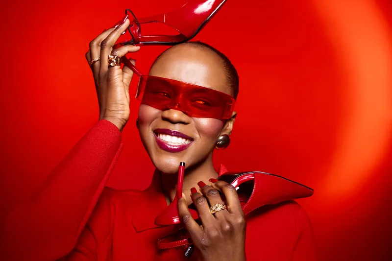Smiling black woman with dark skin wearing red sunglasses and red outfit holds red high heels near face. Bright red background creates vibrant fashion concept with confident model.
