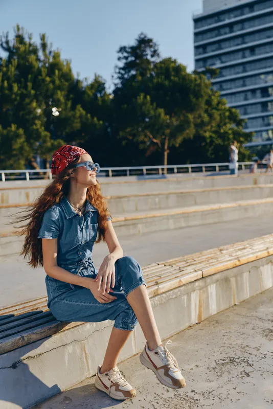 lifestyle woman in modern boho-western denim street style sits relaxed outdoors wearing casual outfit and red bandana in warm film color tones