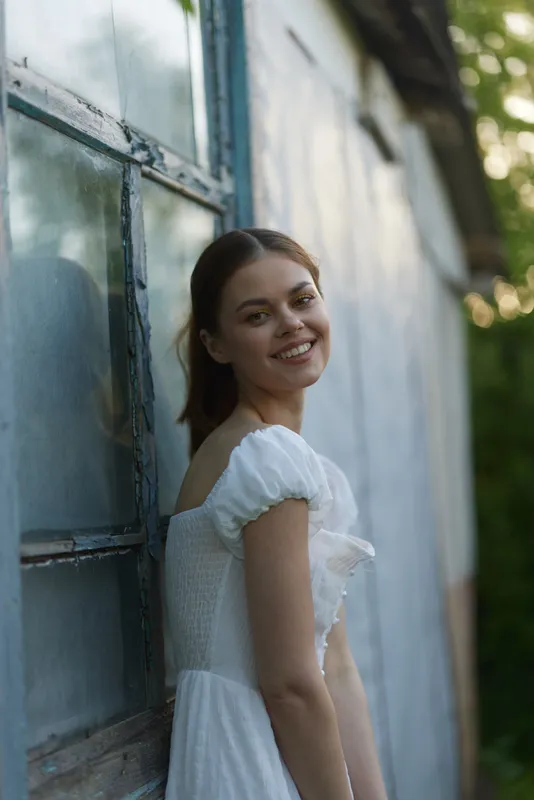 Young woman in a white dress smiling by an old weathered wall, showcasing joy and tranquility in a serene outdoor setting with a soft color palette