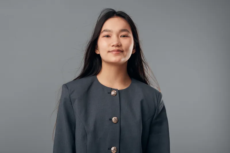 Young woman with long black hair and gray jacket posing with confident smile in front of gray background. Portrait of female model with natural look and casual style indoors.