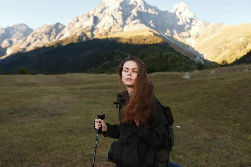 A young female hiker stands in a wide alpine meadow with a trekking pole, framed by rugged mountains. She looks calm and focused, ready for an outdoor adventure.