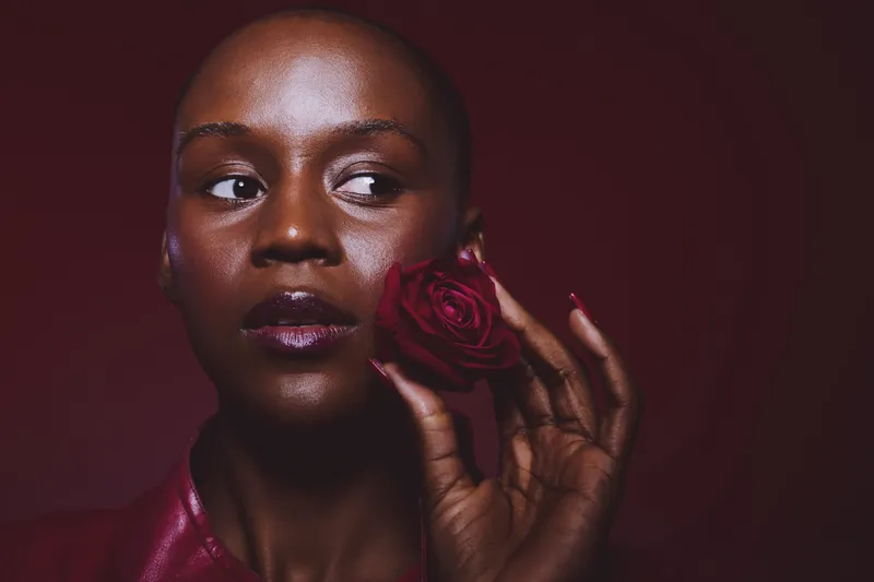 Close-up of a dark skinned African woman with bald head holding a red rose near her face. The portrait features glossy lips and smooth skin on a dark red background. Concept of beauty and elegance.