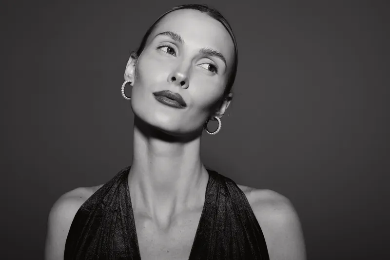 Portrait of a young white woman with pale skin and slicked back hair wearing hoop earrings and a black dress looking away on dark background.