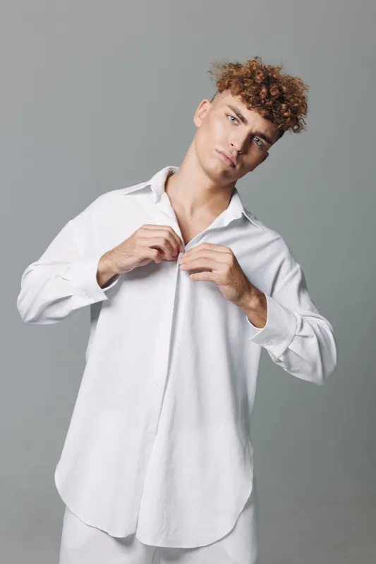 Young man with curly hair wearing oversized white shirt buttoning it up against plain gray background. Casual fashion, studio portrait, confident expression and modern style for lifestyle.