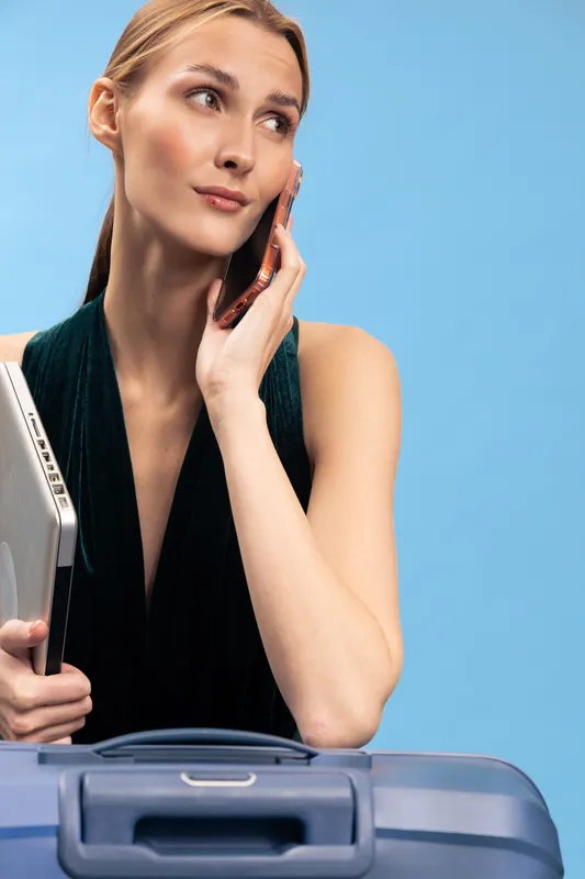 Young woman holding laptop and talking on phone looks thoughtful, standing near suitcase on blue background. Concept for travel planning, business trip, and modern lifestyle.