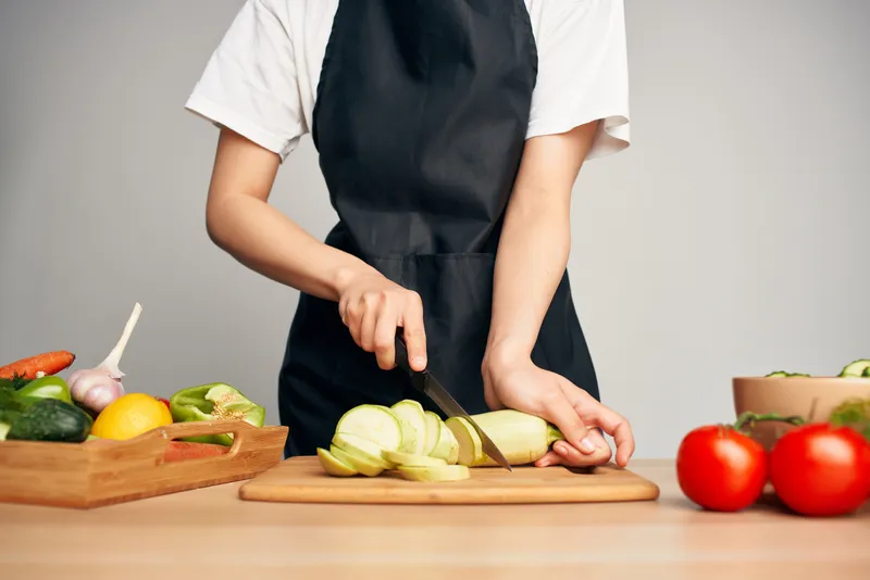 Женщина в черном фартуке нарезает овощи на кухне, готовит еду