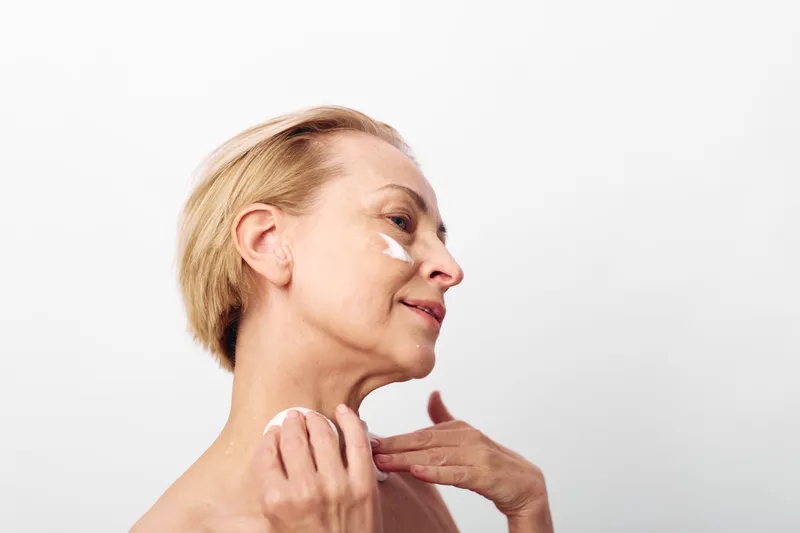 Mature woman with short blonde hair applying face cream on cheeks and neck. Skincare routine concept with natural beauty and self-care on white background in soft lighting.