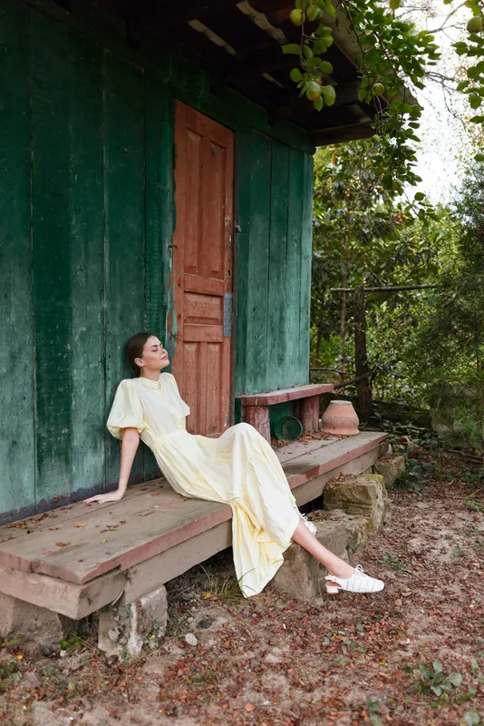 woman in yellow dress on rustic porch bench at green cottage rural relaxation nature scene with authenticity inclusivity peaceful pose vintage summer lifestyle quiet sunlight