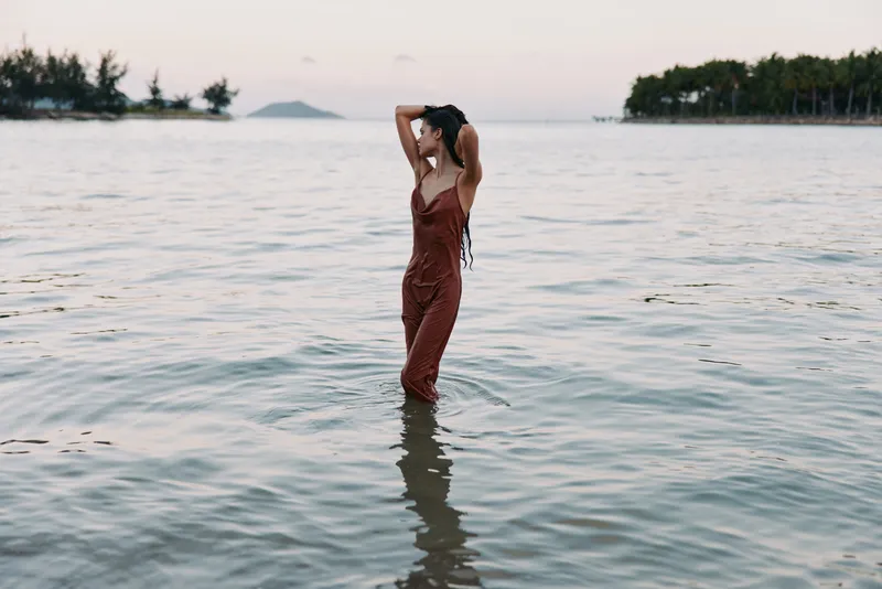Solitary figure stands waist deep in calm coastal water, wearing a rust colored jumpsuit, captured from behind as gentle waves lap the shoreline at dusk.