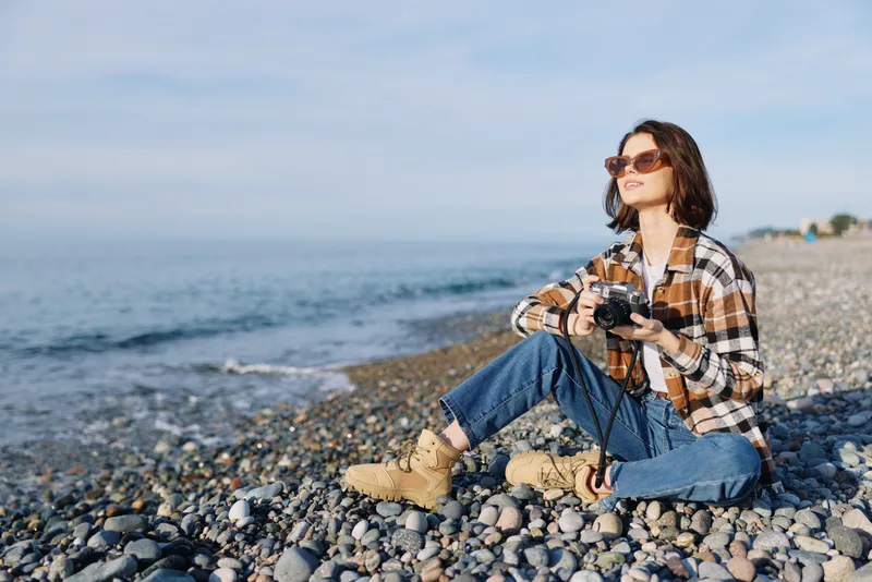Woman photographer sits on a pebbled beach wearing sunglasses and a plaid shirt, holding a camera while enjoying the sea breeze and distant horizon.
