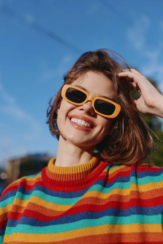Young woman smiling in yellow sunglasses and a colorful striped sweater outdoors on a sunny day, short brown hair and hand touching hair in an urban park under blue sky and natural light.