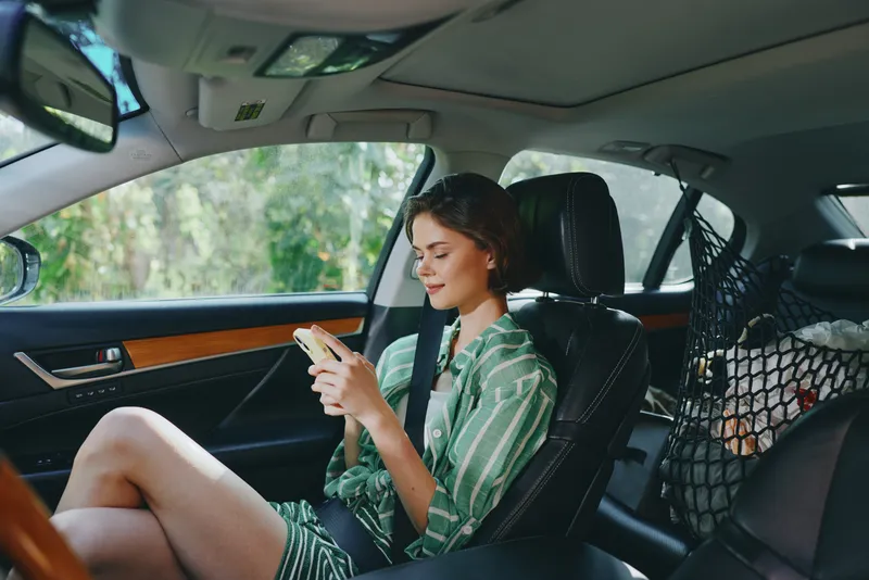 A young woman sits in a car seat and uses a smartphone, relaxed and focused, with sunlight on her face and a tidy cabin.