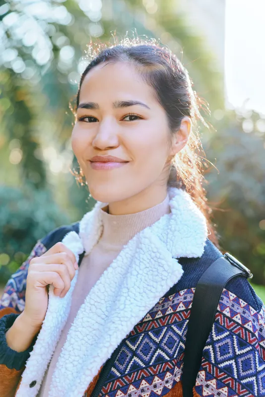 Smiling woman outdoors with backpack and cozy patterned jacket, enjoying a sunny day in a leafy setting, ready for travel, exploration and outdoor activity with relaxed confidence and casual style