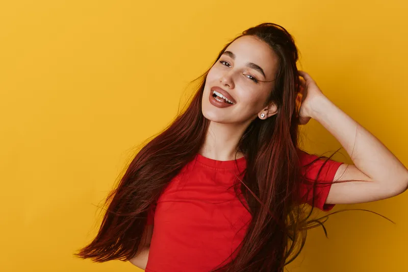 Young woman with long hair smiling and posing in red t shirt on yellow background. Happy female model with natural makeup and earrings expressing joy and confidence.